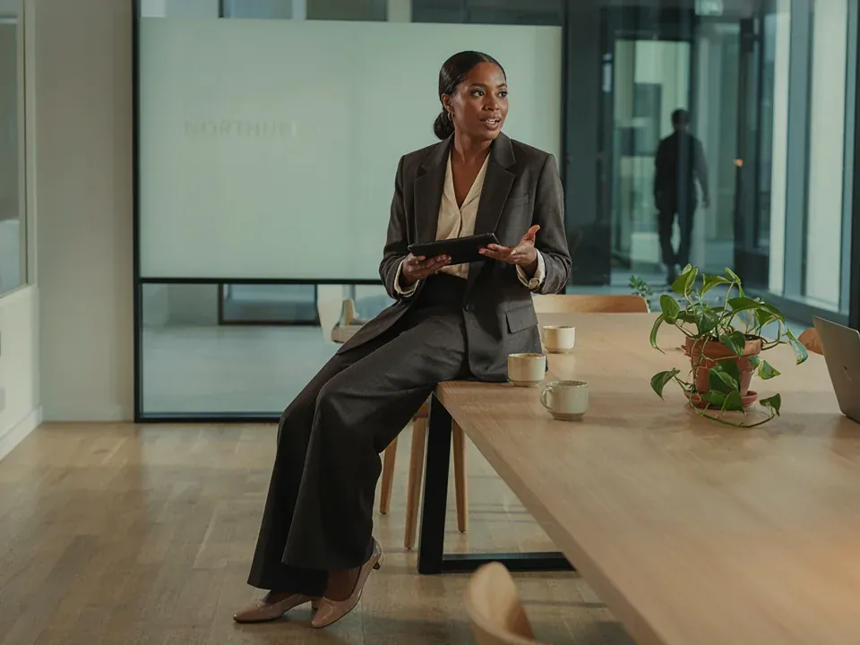 Black businesswoman in a suit, tablet in hand, leading a discussion around a large table in an upscale coworking space