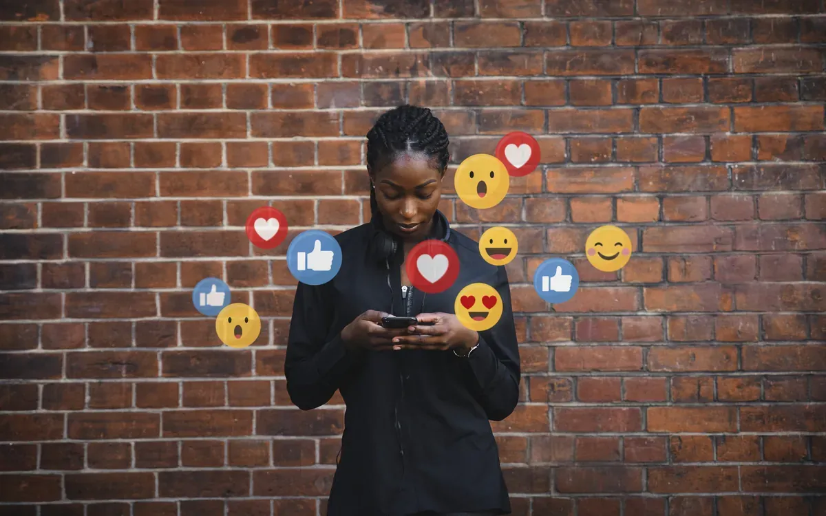 Jeune femme noire consultant son smartphone devant un mur de briques, entourée d'icônes de réactions des réseaux sociaux